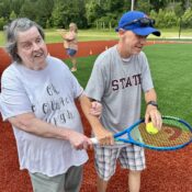 Kamassa camper carrying a tennis racquet and ball with helper on the baseball field at Camp Kamassa.